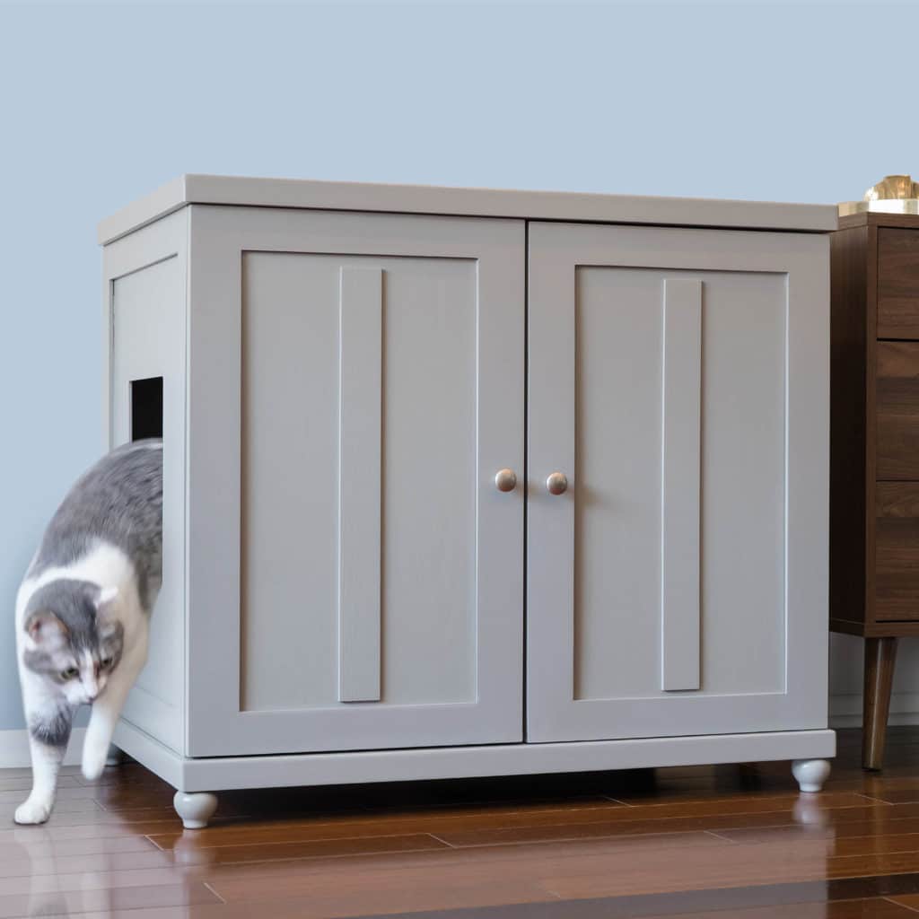 A gray cat leaves a gray wooden cabinet litter box with double doors on a wooden floor next to brown furniture.