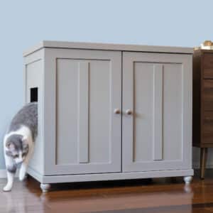 A gray cat leaves a gray wooden cabinet litter box with double doors on a wooden floor next to brown furniture.