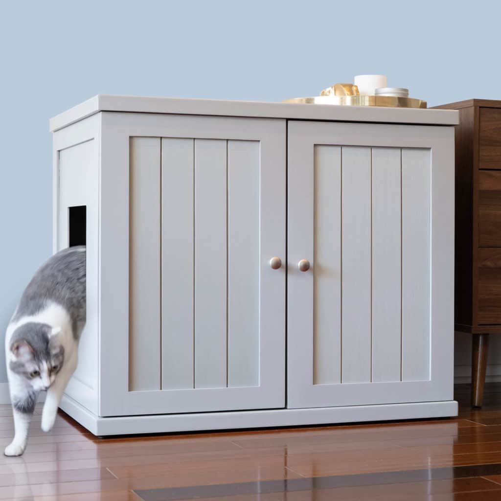 Gray and white cat leaving a white wooden litter box with side entry on wooden floor next to a brown table.