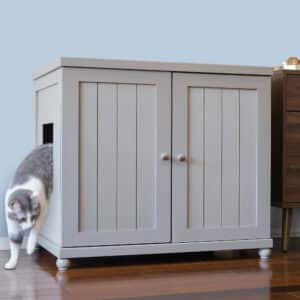 A gray and white cat steps out of a light gray wooden litter box cabinet on a hardwood floor.