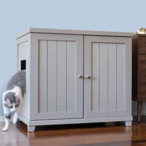 Gray and white cat exiting from a side opening of a double-door litter box cabinet on a wooden floor.