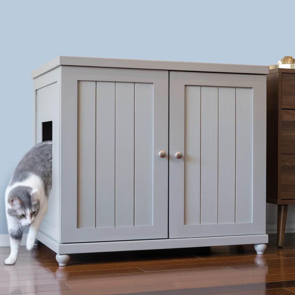 Gray and white cat emerging from side opening of light gray litter box cabinet with double doors on hardwood floor.
