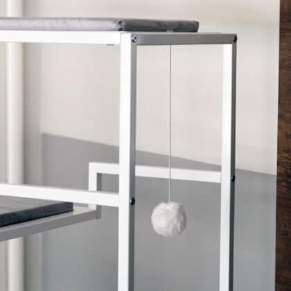 A white metal-framed cat lounge with a gray cushion and a white fluffy ball hanging on a string for play.