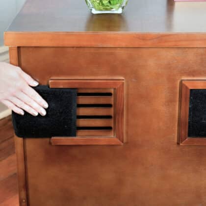 A hand inserts a Charcoal Litter Box Filter into a vent located on the side panel of a wooden cabinet.