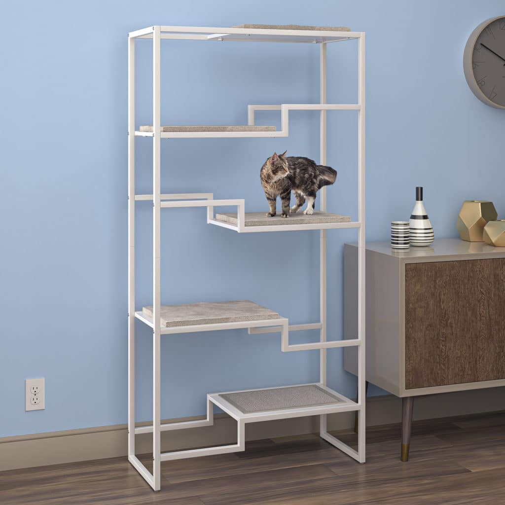 A cat stands on the second shelf of a white-framed, gray-cushioned condo near a wooden cabinet and blue wall.