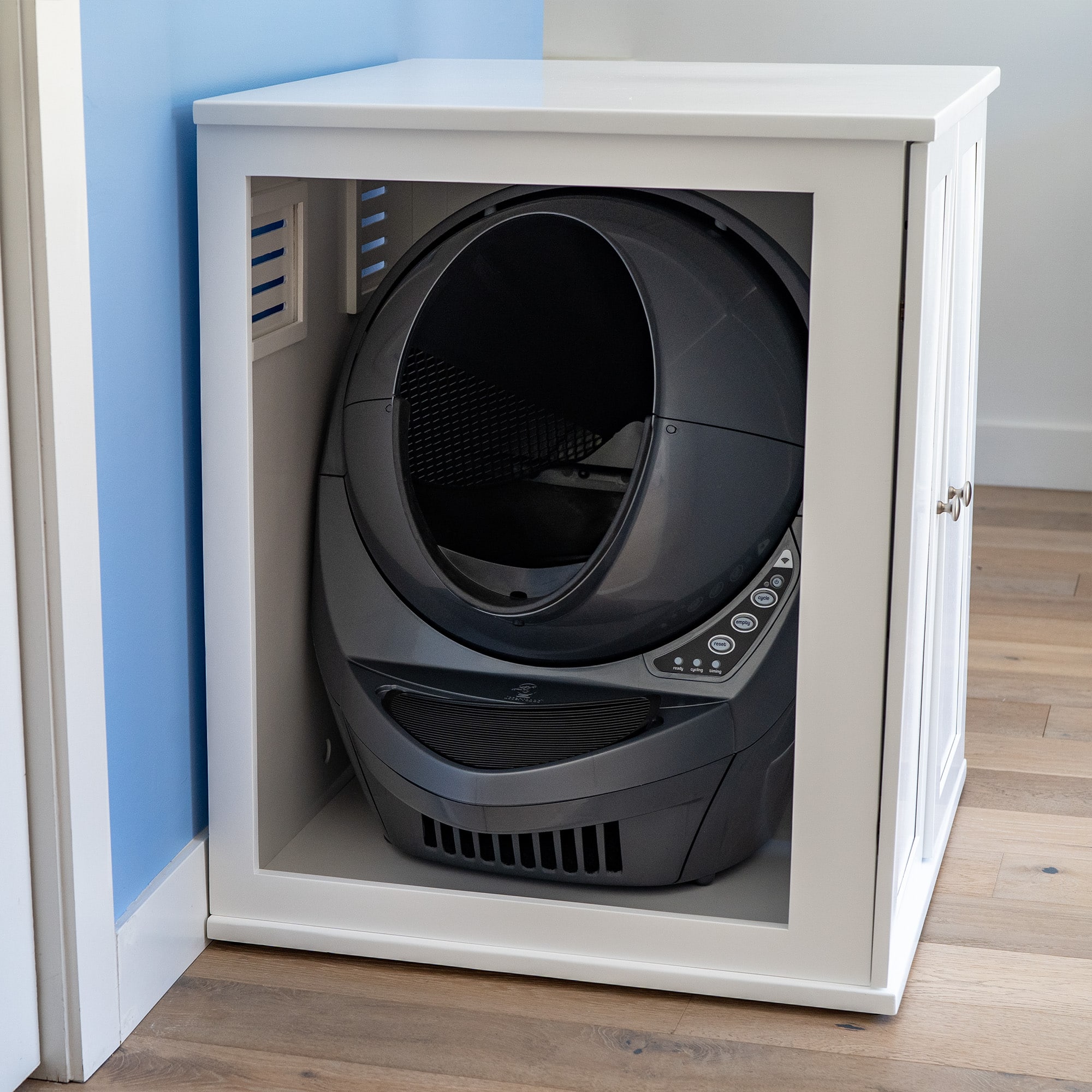 A black self-cleaning litter box inside a white cabinet with a door, on wooden flooring by a light blue and white wall.