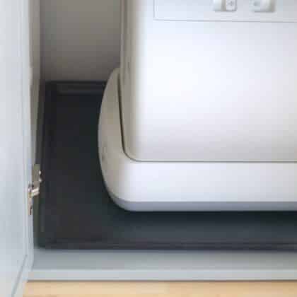 White self-cleaning litter box on a protective tray, placed over a black mat inside an open cabinet, close-up view.