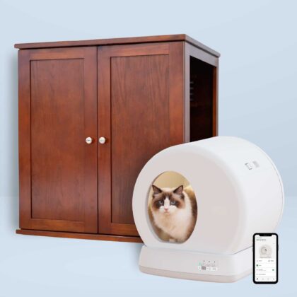 Cat sitting in a white self-cleaning litter box near a wooden cabinet, with a smartphone showing an app beside it.