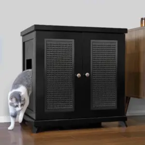 Gray and white cat exits the side opening of a black mesh-door cabinet on a hardwood floor beside brown furniture.