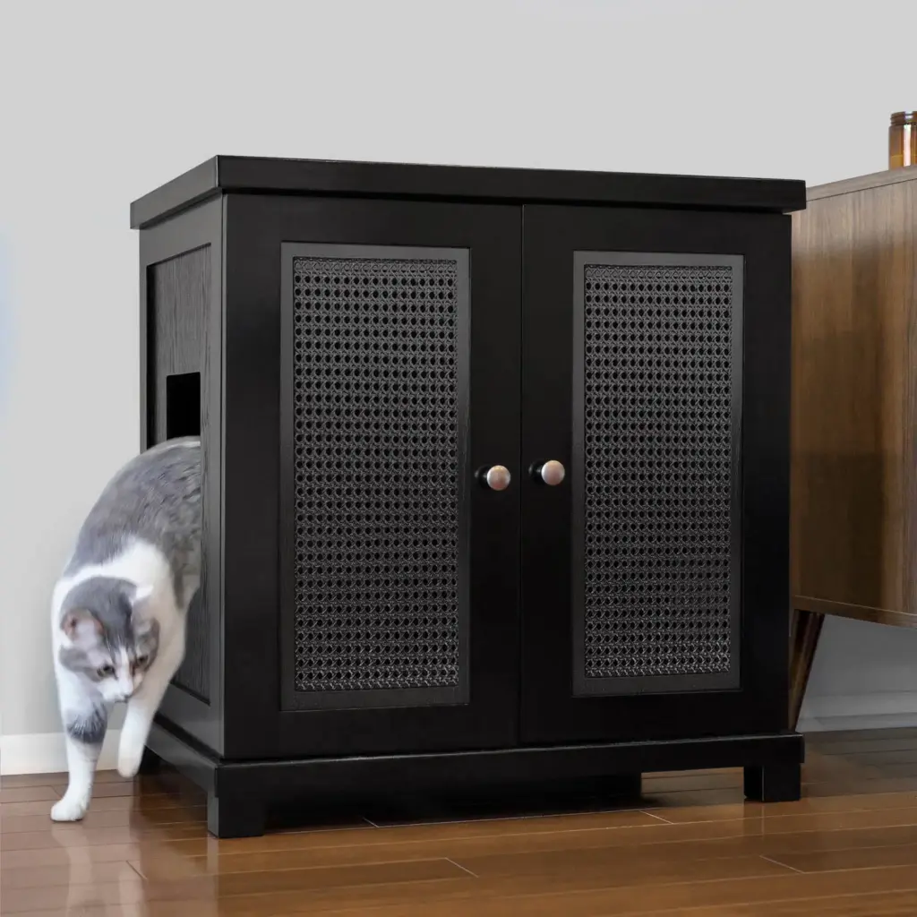 A gray and white cat exits a side opening of a black cabinet with double doors and paneling on a wooden floor.