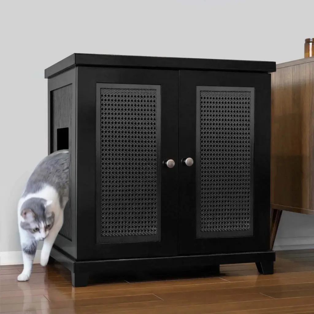 A gray and white cat exits a black wooden cabinet with a side entry hole, on a wood floor beside another furniture piece.