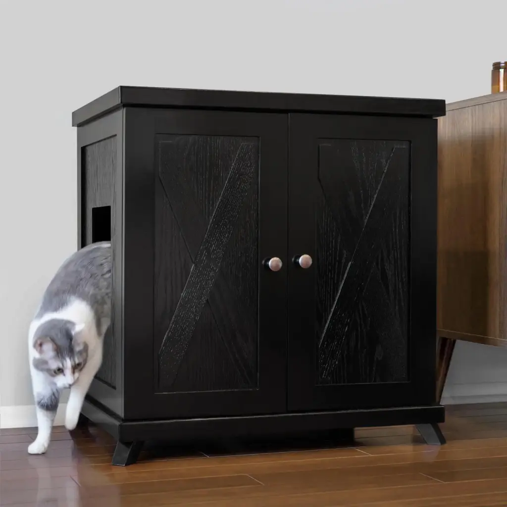 Gray and white cat leaves a black wooden cabinet with a side door on a wooden floor next to another piece of furniture.