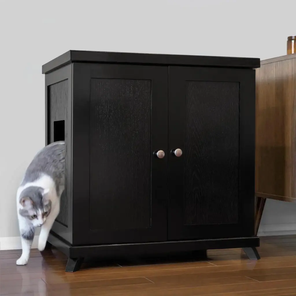 Gray and white cat walks out of a black wooden cabinet with side opening, on hardwood floor next to another furniture piece.