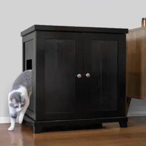 Gray and white cat exiting a black wooden cabinet with a side entry hole, on a wooden floor beside other furniture.
