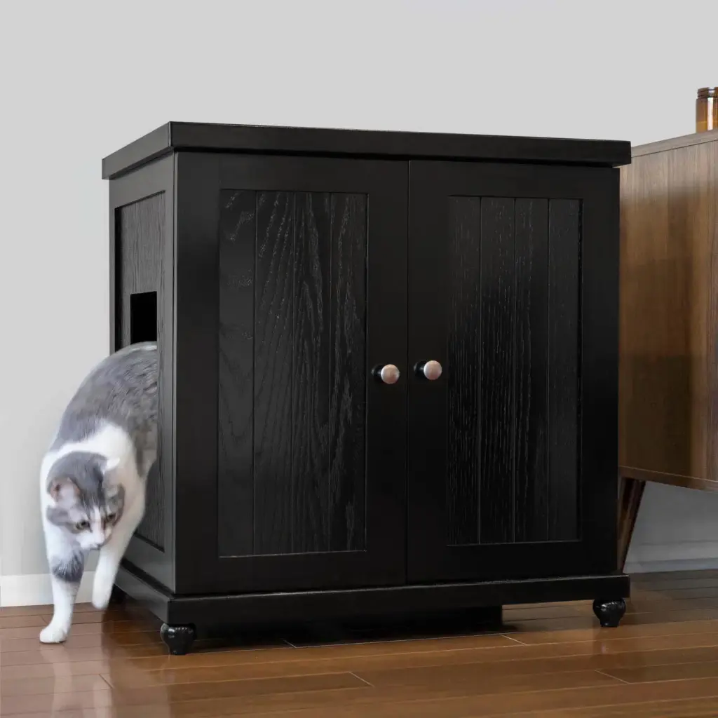 Gray and white cat exits a black wooden cabinet with a side opening, on a wooden floor beside another cabinet.