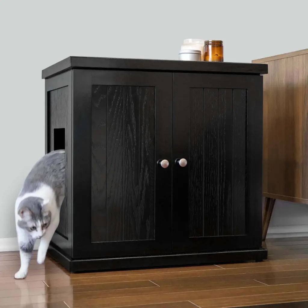 A cat steps out of a black cabinet with a side entrance; two jars are on top, on hardwood floor beside another cabinet.
