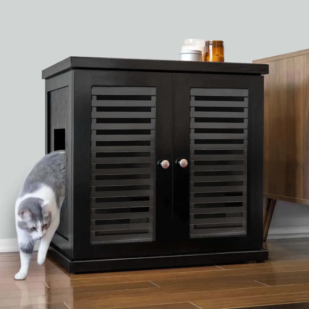 Gray and white cat steps out of a black wooden cabinet with slatted doors; two jars are on top, on hardwood floor.