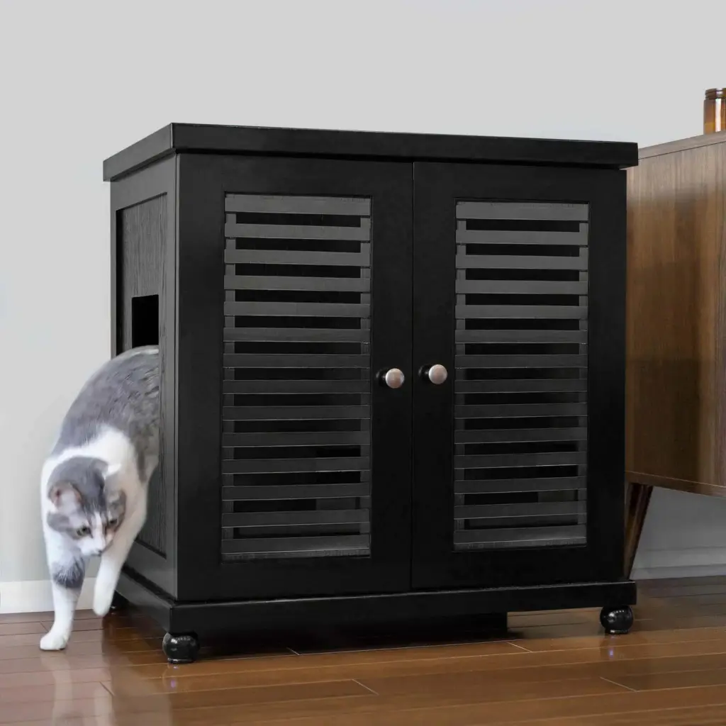 Gray and white cat exits a black wooden cabinet with slatted doors on a hardwood floor beside brown furniture.