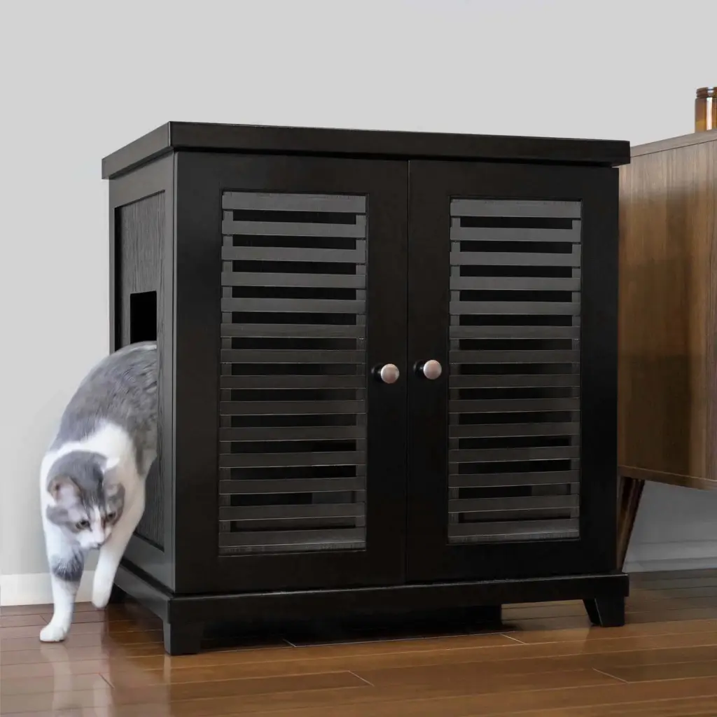 Gray and white cat walks out of a black cabinet with louvered doors onto hardwood floor next to a wooden table.