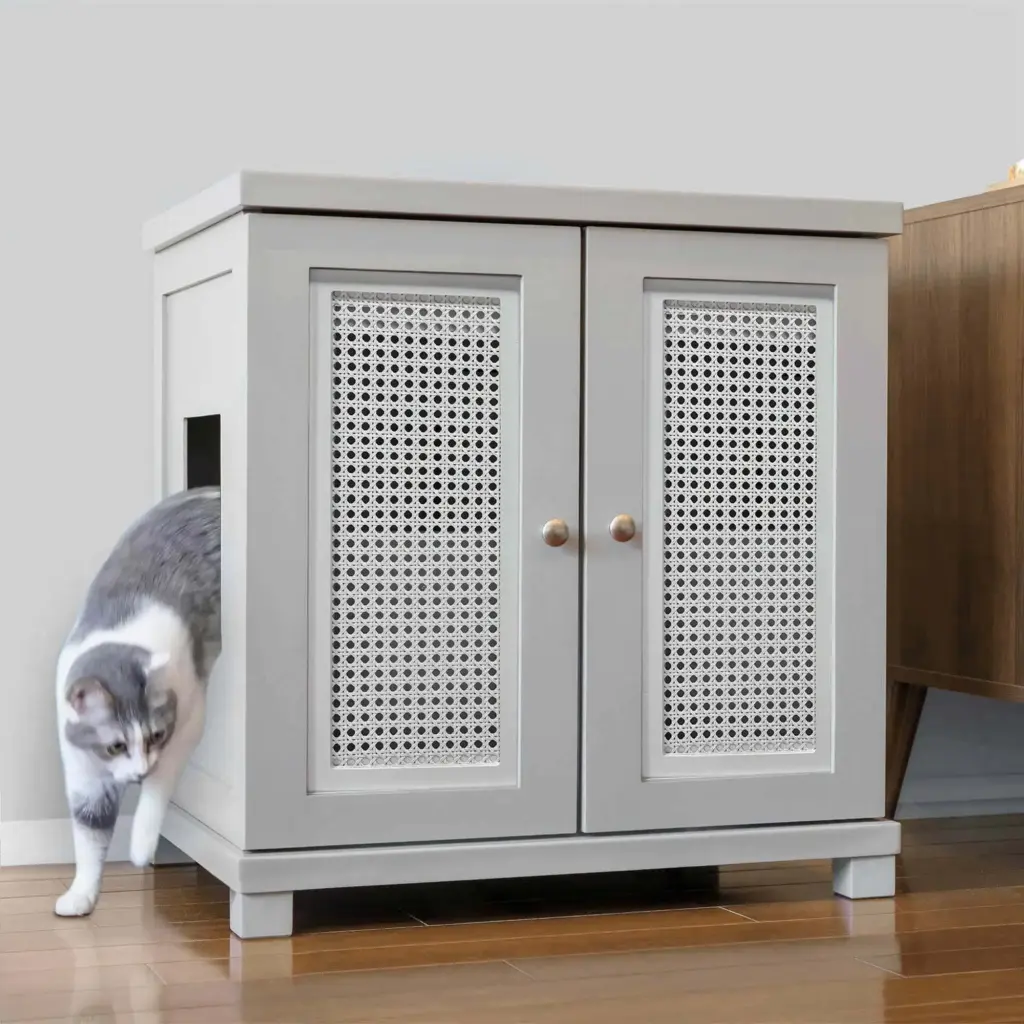 A gray and white cat steps out of a light gray, two-door cabinet with decorative panels on a wooden floor.