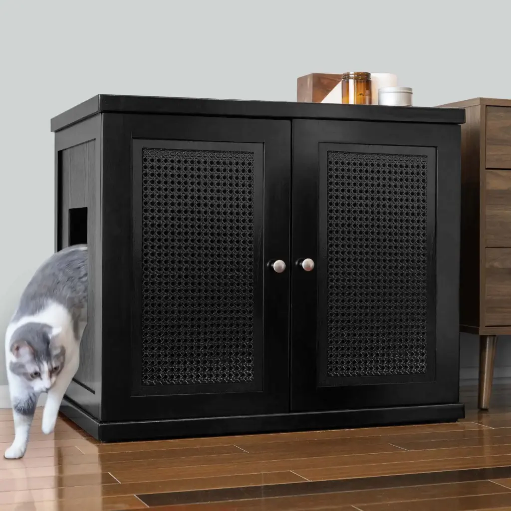 Gray and white cat leaving a black cabinet with lattice doors on hardwood floor next to a brown side table.
