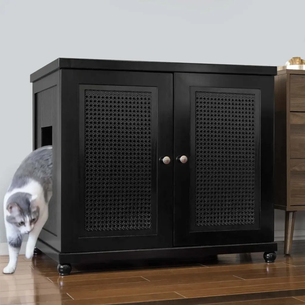 A gray and white cat leaves a black cabinet with a side opening on a wooden floor next to another piece of furniture.