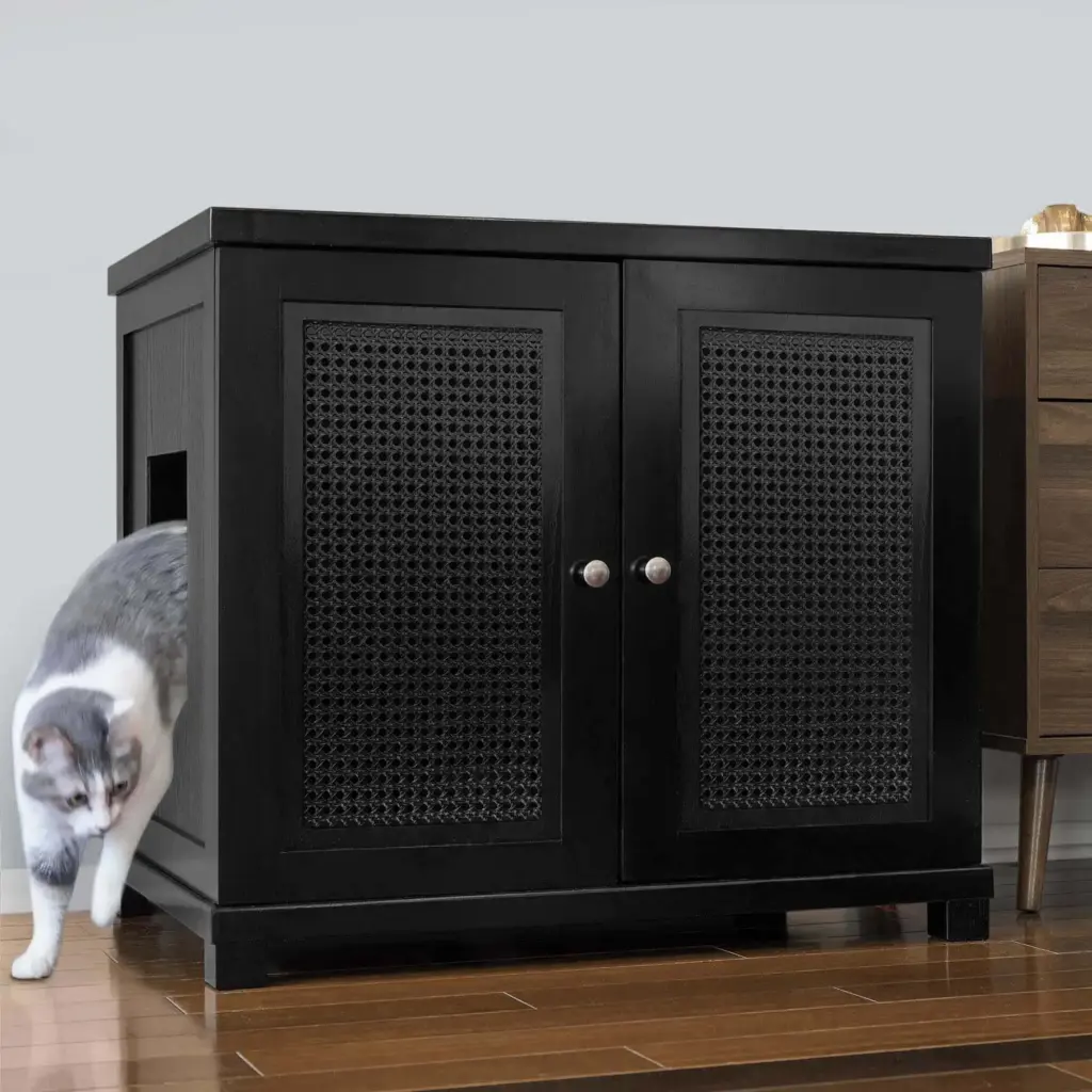Gray and white cat leaving a black cabinet with mesh panel and cutout door on wooden floor beside another furniture piece.