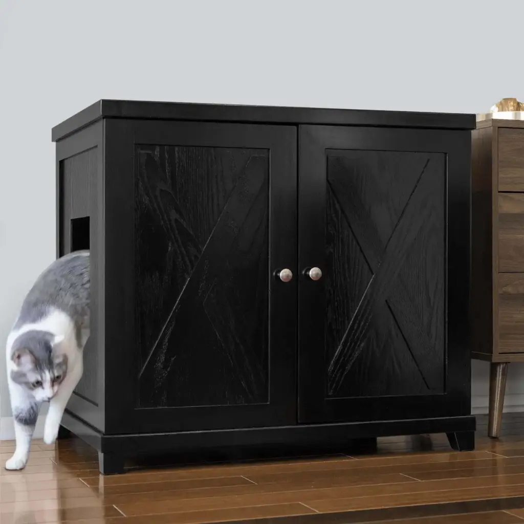 Gray and white cat exits side opening of black double-door cabinet on wooden floor next to brown furniture.
