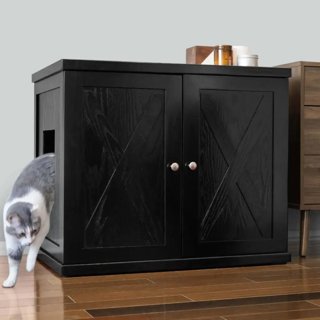 Gray and white cat leaving a black double-door cabinet with side opening on wooden floor beside another piece of furniture.