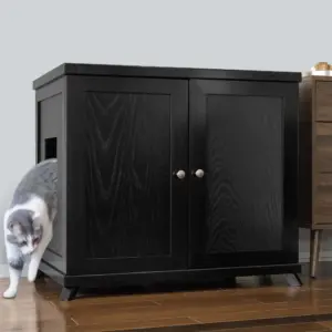 A gray and white cat exits a black wooden cabinet with side entry on a wooden floor beside a brown dresser.