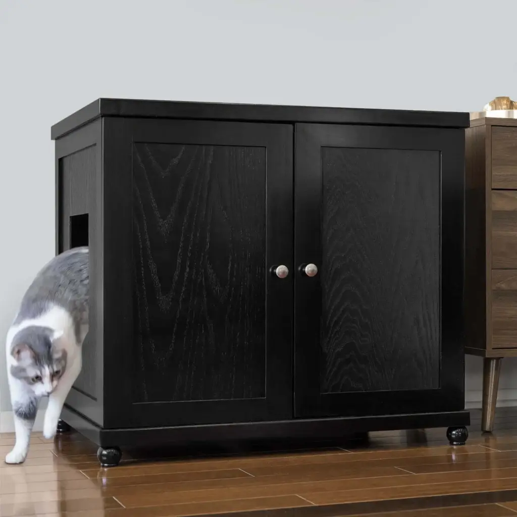 Gray and white cat steps out of a black cabinet with a side opening onto hardwood floor next to another furniture piece.