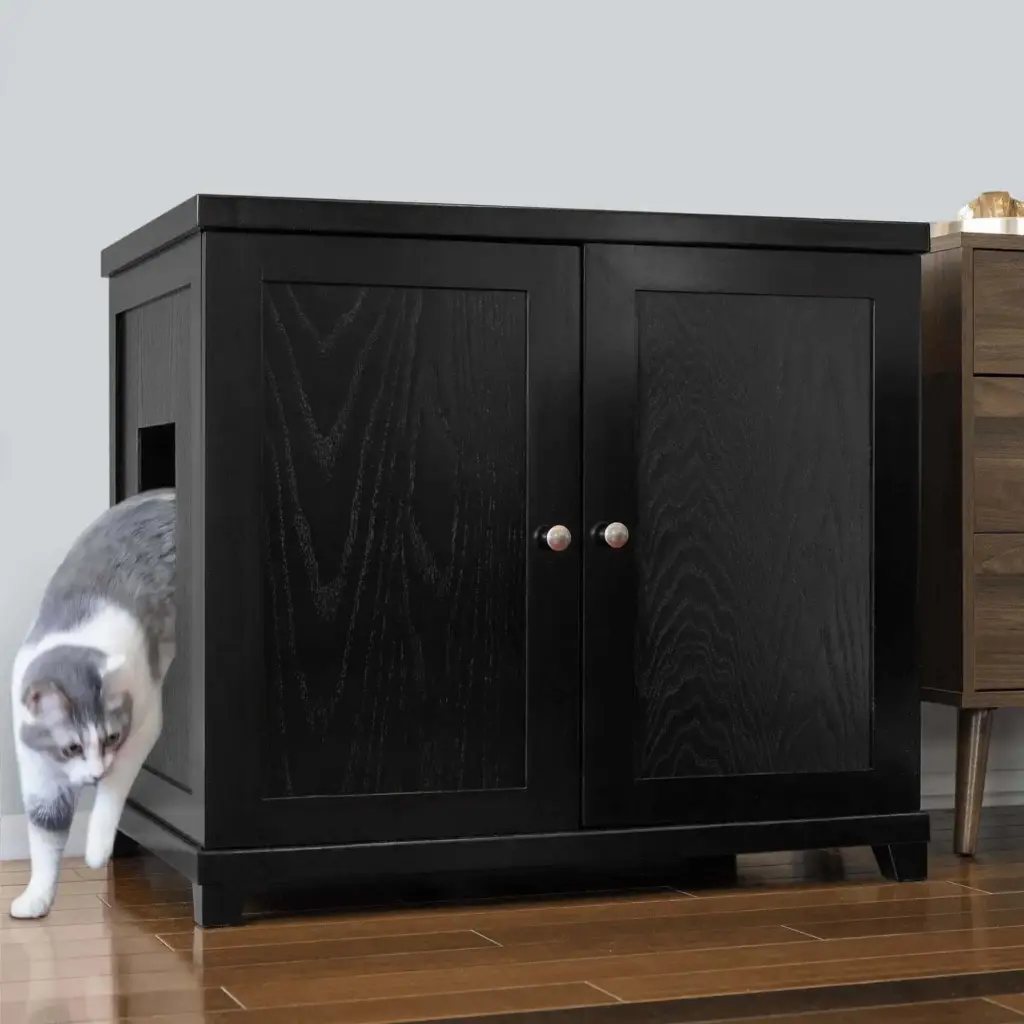 Gray and white cat exiting a black wooden cabinet with side hole, on wood floor beside a brown side table.