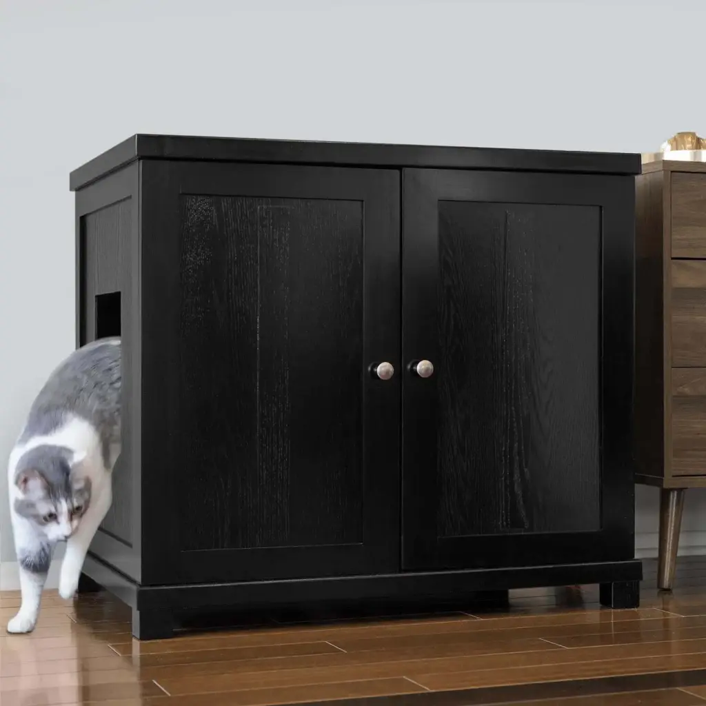 Gray and white cat exiting a black wooden cabinet with a side opening, on wooden floor next to a brown side table.