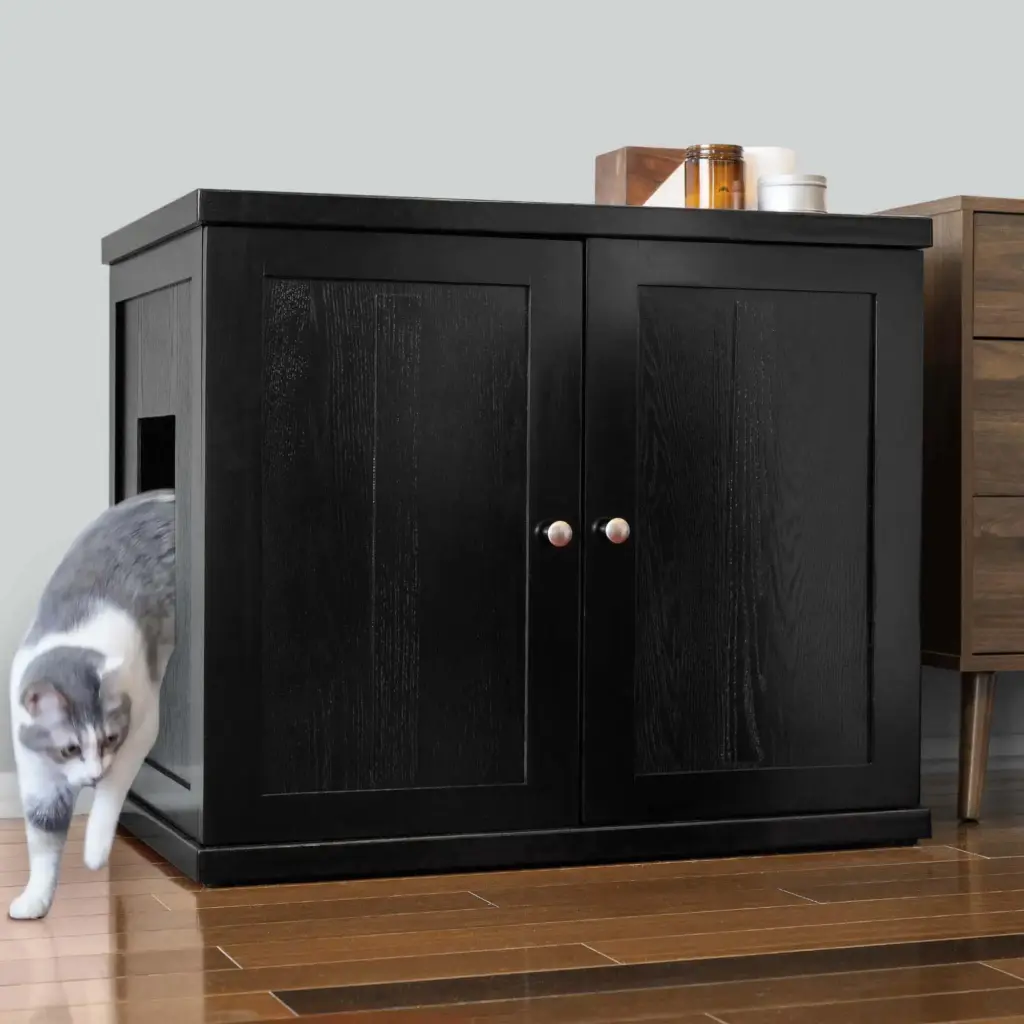 Gray and white cat steps out of a black wooden cabinet with side opening, on brown wood floor next to other furniture.