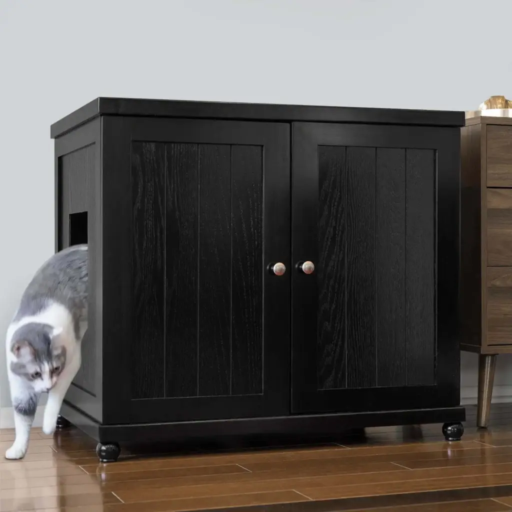 Gray and white cat exits side opening of black cabinet with double doors on wood floor beside small brown dresser.