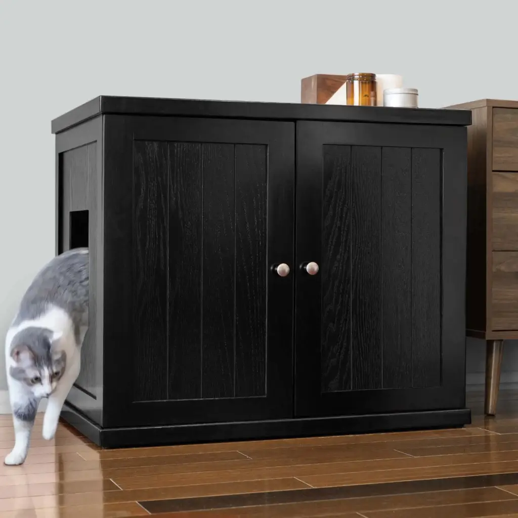 Gray and white cat steps out of a black cabinet with a side opening, near wood floor and small table holding candles.