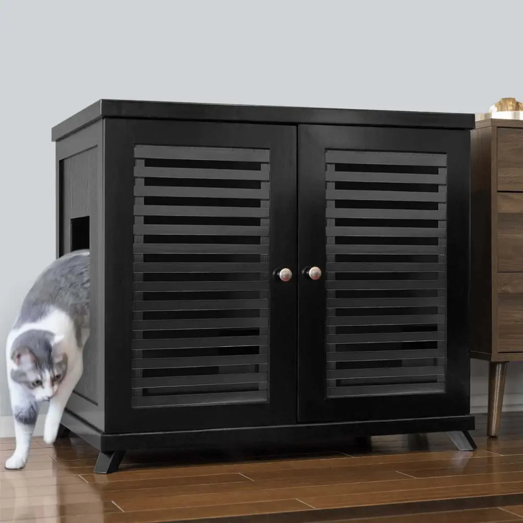 A gray and white cat steps out of a black cabinet with slatted doors on a hardwood floor beside another piece of furniture.