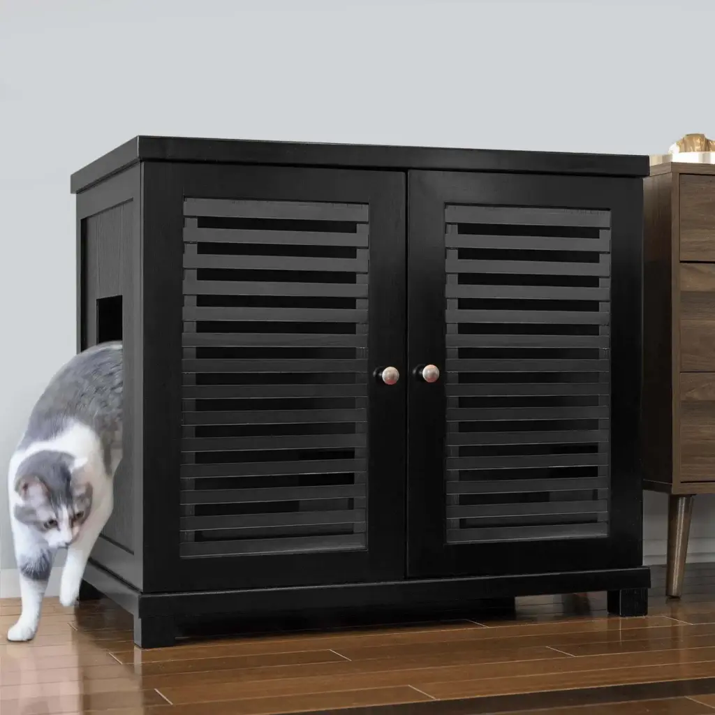 Gray and white cat steps out of a black vented cabinet with side opening on wooden floor beside other furniture.