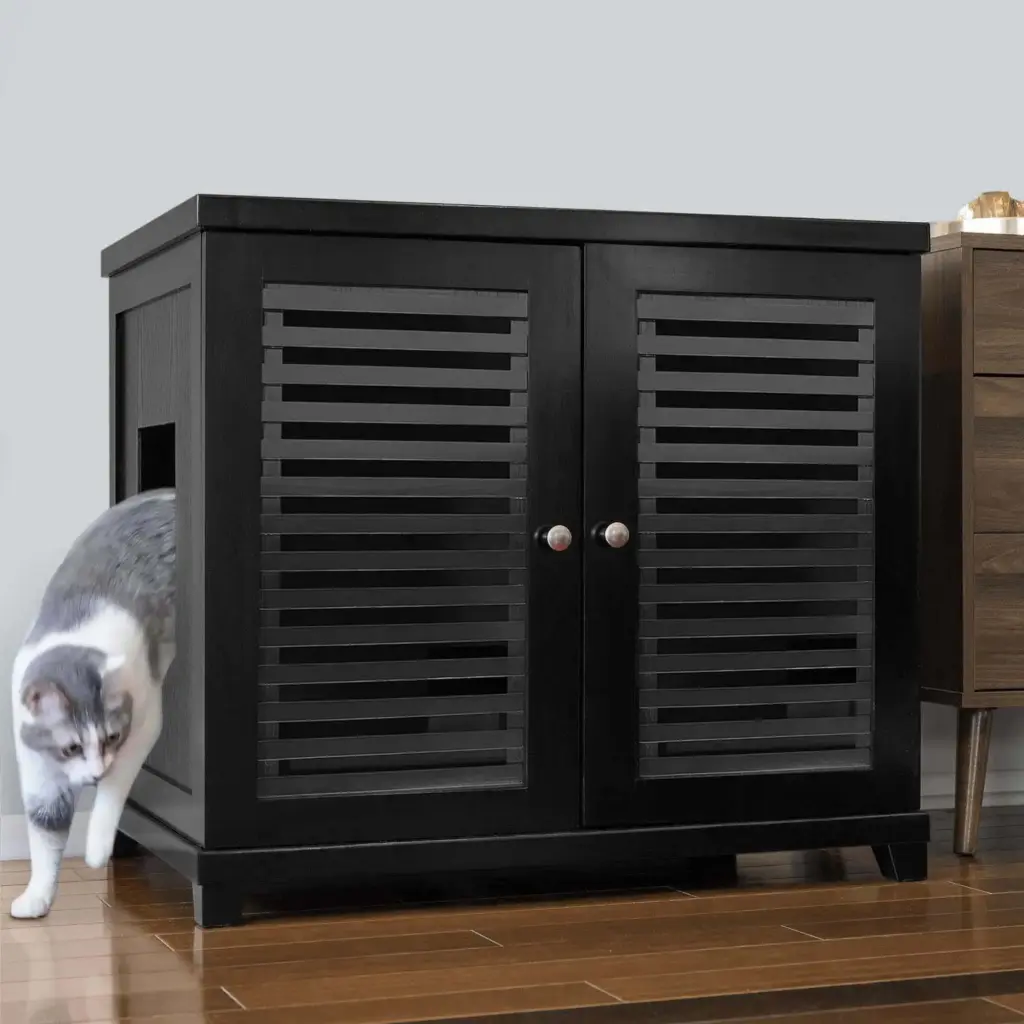 Gray and white cat walking out of a black cabinet with slatted doors on a wooden floor next to other furniture.