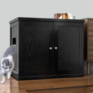 A gray and white cat walks out of a black wooden cabinet with a side opening on a wooden floor beside brown furniture.
