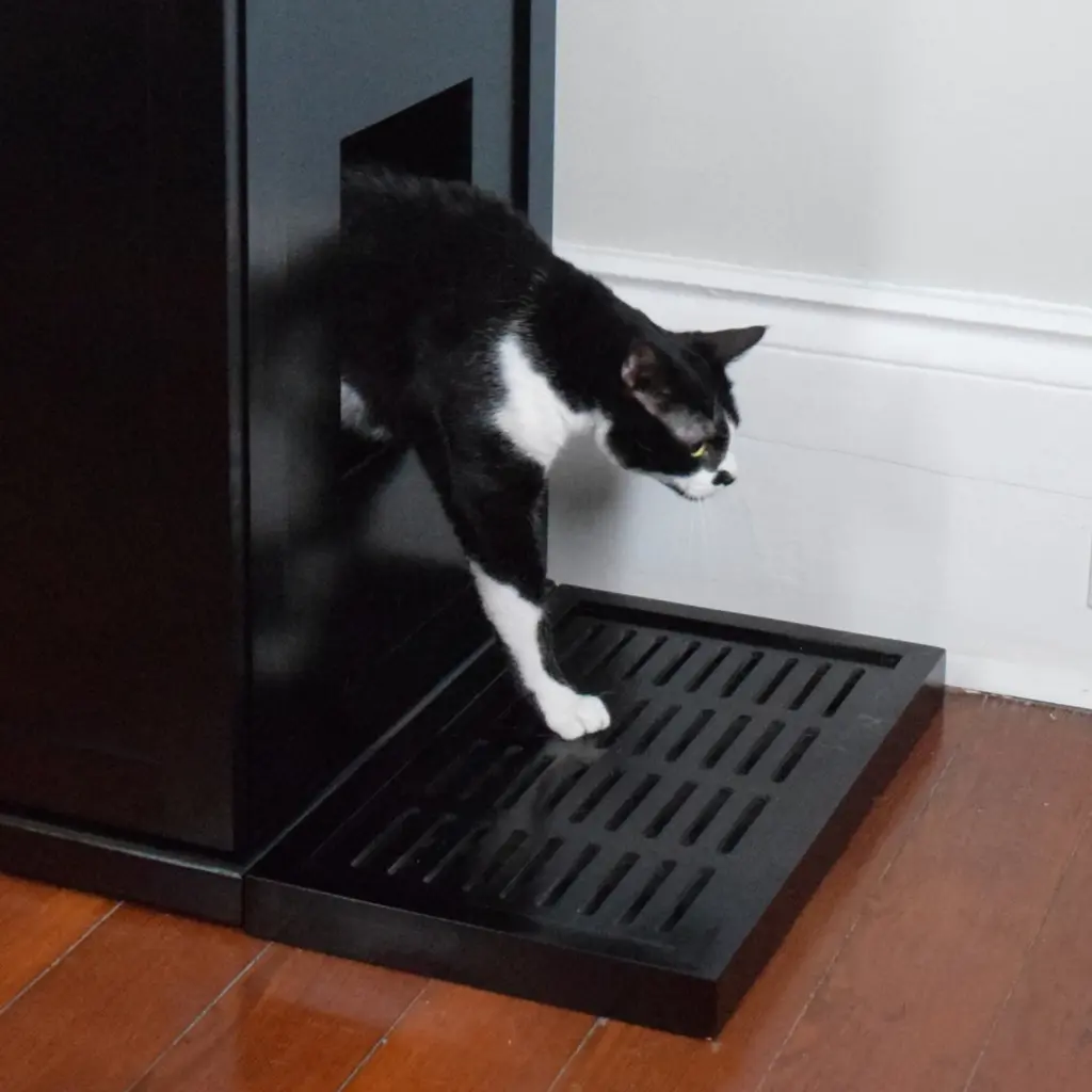A black and white cat steps from a black litter box cabinet onto an ebony slatted mat on a wooden floor by a white wall.